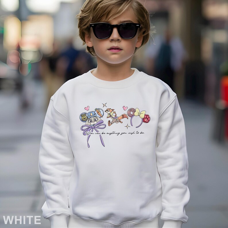Puede incluir: Sudadera blanca con un gr&aacute;fico de dibujos animados coloridos y el texto "You can be anything you wish to be." El dise&ntilde;o incluye un lazo, corazones y diademas tem&aacute;ticas de personajes. La sudadera la lleva una persona con gafas de sol.