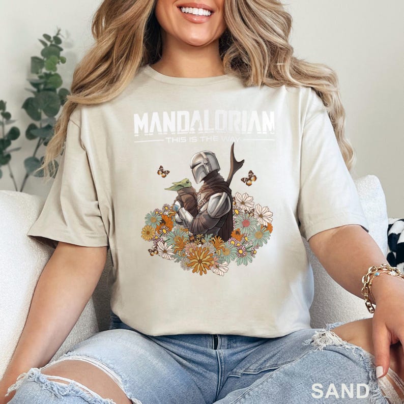 May include: A beige t-shirt with a graphic print of a Mandalorian character holding a small green creature. The text "Mandalorian - This is the way" is printed above the graphic. The graphic is surrounded by flowers and butterflies.