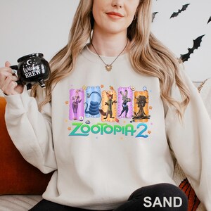 May include: Sand-colored sweatshirt featuring a colorful graphic of characters from the movie "Zootopia 2". The design includes the text "Zootopia 2" in a vibrant, multi-colored font. The sweatshirt is a casual, comfortable style.