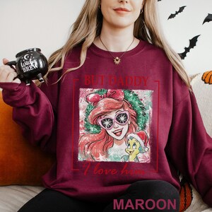 Puede incluir: Sudadera granate con un gr&aacute;fico de dibujos animados de un personaje con gafas de sol rosas y el texto "BUT DADDY I love him". La palabra "MAROON" est&aacute; impresa en la parte inferior de la sudadera.