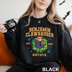 Op de afbeelding: Zwarte sweatshirt met Benjamin Clawhauser uit Zootopia. Het ontwerp bevat de tekst "BENJAMIN CLAWHAUSER" boven een illustratie van het personage in een krans, met "EST. 2016" en "ZOOTOPIA" eronder. Het woord "BLACK" staat onderaan.