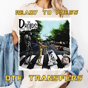 May include: A white t-shirt featuring a graphic of Star Wars droids crossing a street, with the text "The Droids Imperial Road." The words "Ready to Press" and "DTF Transfers" are also displayed in gold.