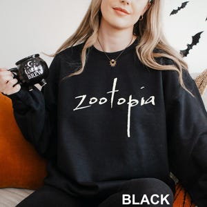 May include: Black sweatshirt featuring the word "zootopia" in white script. The word "BLACK" is printed in white block letters at the bottom. A black mug with "Witches Brew" is held in the left hand. Halloween theme.