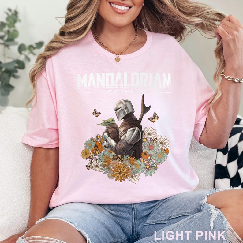 May include: A light pink t-shirt with a white graphic of the Mandalorian and Grogu surrounded by flowers and butterflies. The text "MANDALORIAN THIS IS THE WAY" is printed on the shirt.
