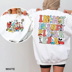 May include: White sweatshirt featuring a colorful graphic of Disney and Pixar characters on the front and back. The back of the sweatshirt has the text "IN MY DISNEY PIXAR ERA" in a retro font. The sweatshirt is made of a soft material.