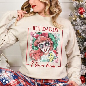 Puede incluir: Sudadera color arena con un gr&aacute;fico de Ariel de La Sirenita con gafas de sol, con el texto "BUT DADDY I love him." El dise&ntilde;o incluye una corona navide&ntilde;a y Flounder con un gorro de Pap&aacute; Noel.