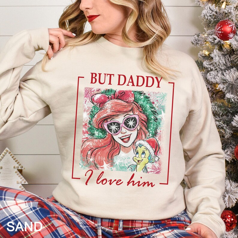 Puede incluir: Sudadera color arena con un gr&aacute;fico de Ariel de La Sirenita con gafas de sol, con el texto "BUT DADDY I love him." El dise&ntilde;o incluye una corona navide&ntilde;a y Flounder con un gorro de Pap&aacute; Noel.