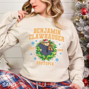 Op de afbeelding: Beige sweatshirt met de oranje tekst "BENJAMIN CLAWHAUSER". Onder de tekst staat een cartoonafbeelding van een personage uit Zootopia met een kerstmuts. De sweatshirt heeft ook de tekst "EST. 2016" en "ZOOTOPIA".