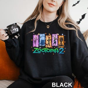 May include: Black sweatshirt featuring a colorful graphic of characters from "Zootopia 2". The design includes the movie title in green and blue, with the word "BLACK" printed at the bottom. The person is holding a black mug that says "Witches Brew".