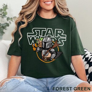 May include: A forest green t-shirt with a white graphic of a cartoon Mandalorian and Grogu from Star Wars. The text "STAR WARS" is printed in white above the characters.