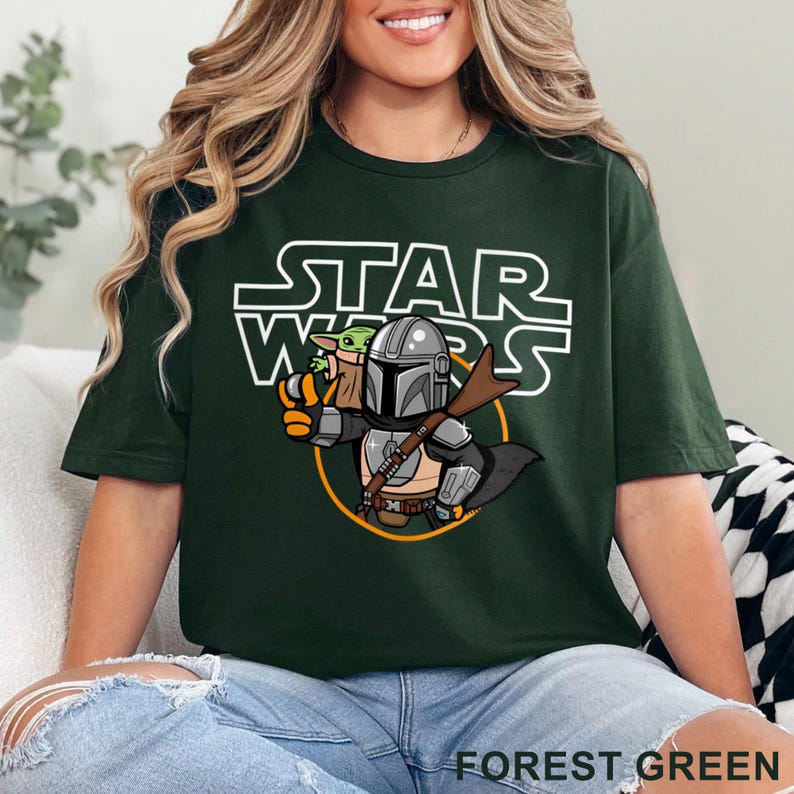 May include: A forest green t-shirt with a white graphic of a cartoon Mandalorian and Grogu from Star Wars. The text "STAR WARS" is printed in white above the characters.