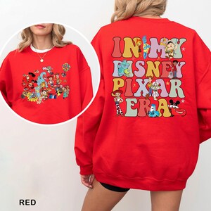 May include: Red sweatshirt featuring a colorful graphic of Disney and Pixar characters. The back of the sweatshirt has the text "IN MY DISNEY PIXAR ERA" in a retro font. The sweatshirt is a crew neck style.