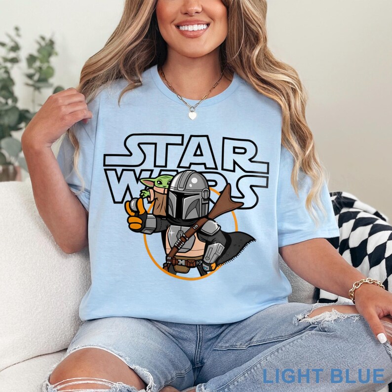 May include: Light blue t-shirt with a cartoon image of a Mandalorian and Grogu from Star Wars. The text "STAR WARS" is printed in black above the image.