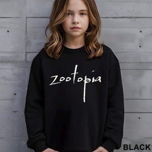 May include: Black crew neck sweatshirt featuring the word "Zootopia" in white script. The sweatshirt is worn by a person. The word "BLACK" is printed in white at the bottom right corner of the image.