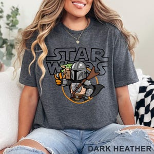 May include: A dark heather grey t-shirt with a cartoon image of the Mandalorian and Grogu from Star Wars. The text "STAR WARS" is printed above the image.