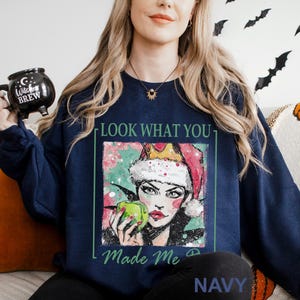 Puede incluir: Sudadera azul marino con un gr&aacute;fico de una mujer con un gorro de Pap&aacute; Noel sosteniendo una manzana verde. El texto "LOOK WHAT YOU Made Me" est&aacute; impreso en la parte delantera. La sudadera tambi&eacute;n tiene la palabra "NAVY" impresa en la parte inferior.