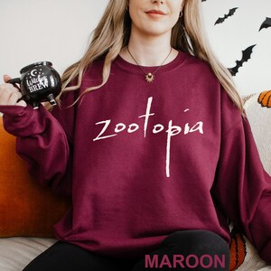 May include: A maroon sweatshirt with the word "Zootopia" in white script. The word "MAROON" is printed at the bottom of the image. A black mug with the words "Witches Brew" is held in the left hand.