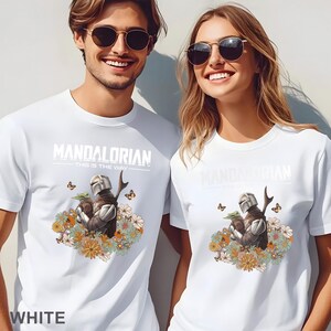 May include: White t-shirt with a graphic of a Mandalorian character holding a baby Yoda surrounded by flowers. The text "Mandalorian - This is the Way -" is printed on the shirt.