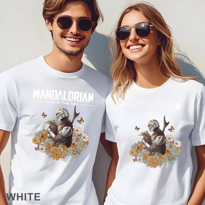 May include: White t-shirt with a graphic of a Mandalorian character holding a baby Yoda surrounded by flowers. The text "Mandalorian - This is the Way -" is printed on the shirt.
