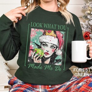 Puede incluir: Sudadera verde bosque con un gr&aacute;fico de una mujer con un gorro de Pap&aacute; Noel y sosteniendo una manzana verde. El texto "LOOK WHAT YOU Made Me Do" est&aacute; impreso en la sudadera. Una taza blanca se sostiene en la mano derecha.