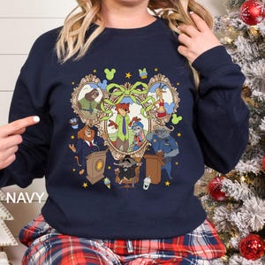 Pode incluir: Camisola azul marinho com uma ilustra&ccedil;&atilde;o de desenho animado colorida de personagens de Zootopia. O design inclui retratos de animais emoldurados, um la&ccedil;o central e detalhes de estrelas. A palavra "NAVY" &eacute; impressa no lado esquerdo da camisola.