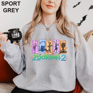 May include: Sport grey sweatshirt featuring a colorful graphic of characters from Zootopia 2. The design includes the text "Zootopia 2" in a vibrant, multi-colored font. The sweatshirt also has the text "SPORT GREY" at the top.