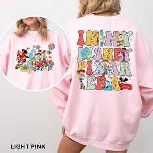 May include: Light pink sweatshirt featuring a colorful graphic of Disney and Pixar characters on the front and back. The back of the sweatshirt says "IN MY DISNEY PIXAR ERA" in a retro font. The front has a collage of characters.