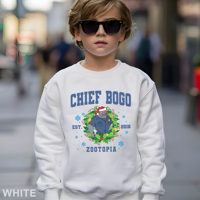 Op de afbeelding: Een witte sweatshirt met de tekst "CHIEF BOGO" in blauw. Daaronder een afbeelding van een gorilla in politie-uniform en kerstmuts, omringd door een krans. De sweatshirt heeft ook "ZOOTOPIA" en "EST. 2016".