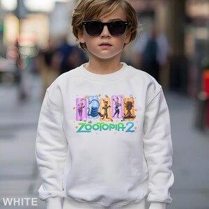 May include: White sweatshirt featuring a colorful graphic of characters from "Zootopia 2". The design includes Judy Hopps, Nick Wilde, and other characters. The text "ZOOTOPIA 2" is printed in a vibrant, multi-colored font. The sweatshirt is a comfortable, casual style.