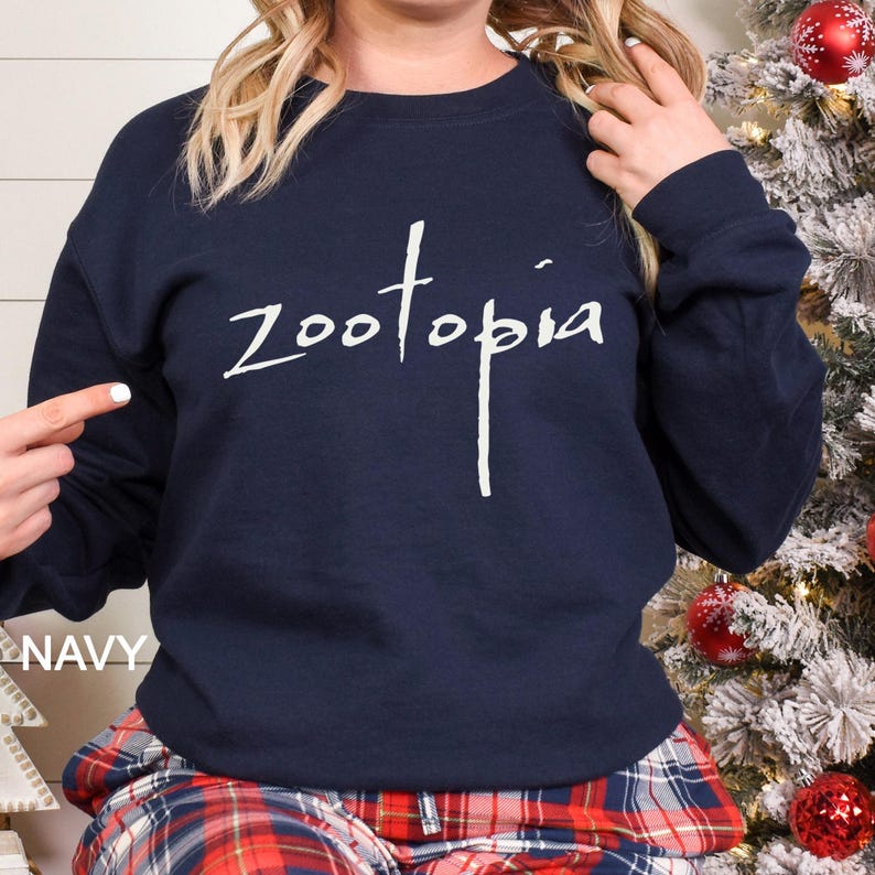 May include: A navy blue crew neck sweatshirt featuring the word "Zootopia" in white script. The word "NAVY" is printed in white at the bottom left of the image. The person is wearing red and blue plaid pyjama bottoms.