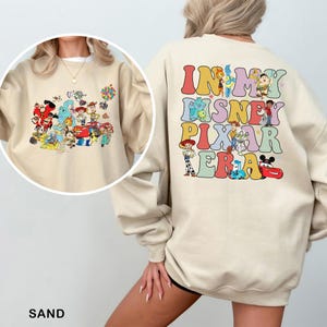 May include: Sand-colored sweatshirt featuring a colorful graphic of Disney Pixar characters on the front and the text "IN MY DISNEY PIXAR ERA" on the back. The sweatshirt has long sleeves and a crew neck.