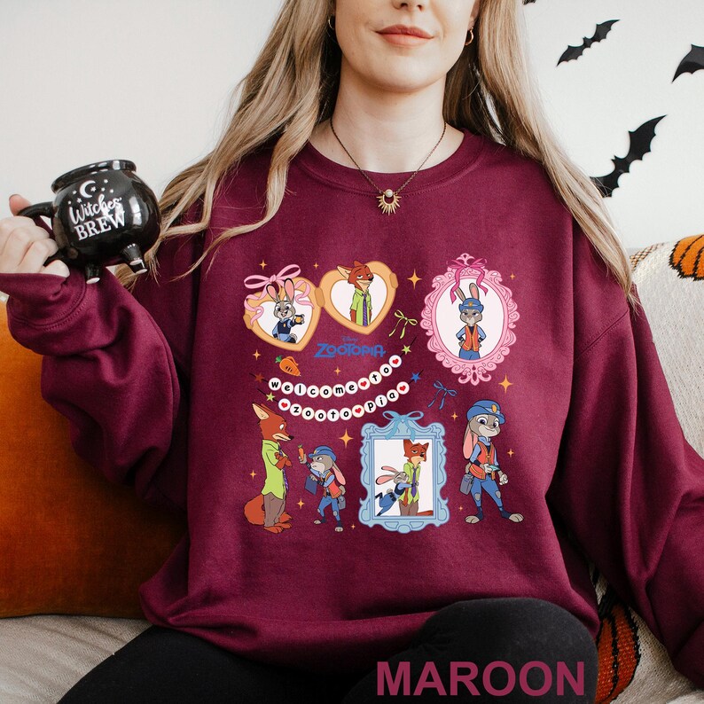 May include: Maroon sweatshirt featuring colorful cartoon illustrations of characters from the movie Zootopia. The design includes framed portraits of a fox and a bunny, along with the word "Zootopia".