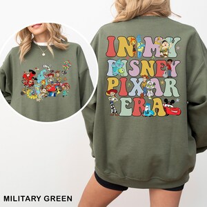 May include: Military green sweatshirt featuring a colorful graphic of Disney Pixar characters on the front and the text "IN MY DISNEY PIXAR ERA" on the back. The sweatshirt has a crew neck and long sleeves, perfect for casual wear.