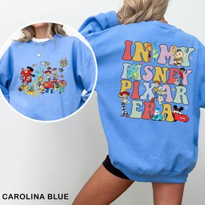 May include: Carolina blue sweatshirt featuring a colorful graphic of Disney and Pixar characters on the front and back. The back of the sweatshirt has the text "IN MY DISNEY PIXAR ERA" in a retro font. The front has a collage of characters.