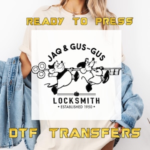 May include: White t-shirt with a black graphic of two cartoon mice carrying a key, with the text "JAQ & GUS-GUS LOCKSMITH ESTABLISHED 1950". The words "READY TO PRESS" and "DTF TRANSFERS" are also displayed.