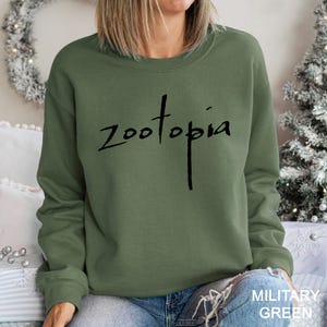 May include: An olive green sweatshirt featuring the word "Zootopia" in black script. The crew neck sweatshirt has a solid colour. The text "MILITARY GREEN" is printed in white at the bottom right.
