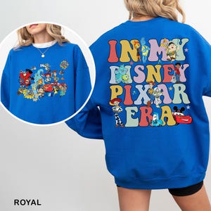 May include: Royal blue sweatshirt featuring a colorful graphic of Disney and Pixar characters on the front and back. The back of the sweatshirt has the text "IN MY DISNEY PIXAR ERA" in a retro font. The sweatshirt is a comfortable, casual style.