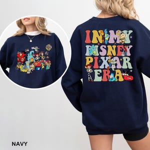 May include: Navy blue sweatshirt featuring a colorful graphic of Disney and Pixar characters on the front and the text "IN MY DISNEY PIXAR ERA" on the back. The sweatshirt has a crew neck and long sleeves.