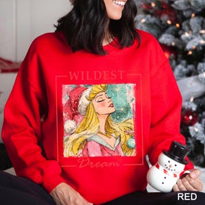 Puede incluir: Una sudadera roja con un gr&aacute;fico de una princesa con un gorro de Pap&aacute; Noel, con las palabras "WILDEST Dream" impresas arriba y abajo de la imagen. La princesa tiene el pelo rubio y sostiene una bola de Navidad.