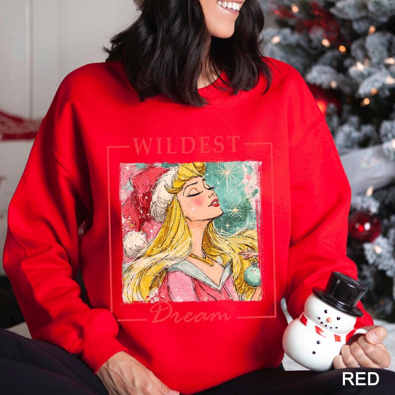 Puede incluir: Una sudadera roja con un gr&aacute;fico de una princesa con un gorro de Pap&aacute; Noel, con las palabras "WILDEST Dream" impresas arriba y abajo de la imagen. La princesa tiene el pelo rubio y sostiene una bola de Navidad.