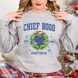 Op de afbeelding: Sportgrijs sweatshirt met de tekst "CHIEF BOGO" en een cartoonafbeelding van Chief Bogo uit Zootopia met een kerstmuts en politie-uniform, omringd door een kerstkrans. De tekst "EST. 2016 ZOOTOPIA" staat er ook op.