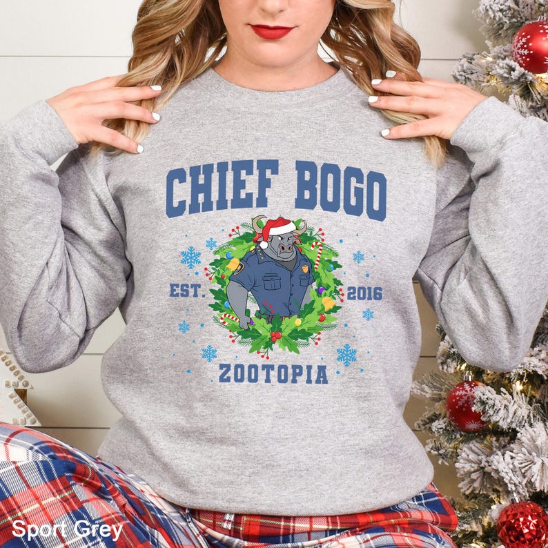 Op de afbeelding: Sportgrijs sweatshirt met de tekst "CHIEF BOGO" en een cartoonafbeelding van Chief Bogo uit Zootopia met een kerstmuts en politie-uniform, omringd door een kerstkrans. De tekst "EST. 2016 ZOOTOPIA" staat er ook op.
