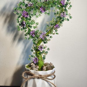 May include: A beaded tree sculpture in a white geometric pot, featuring a heart-shaped crown of green leaves and purple flowers. The tree trunk is green, and the pot is adorned with small rabbit figurines and a jute bow.