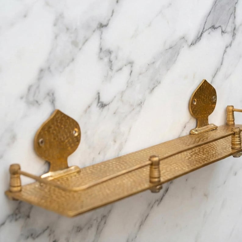 Unlacquered brass floating shelf used as a kitchen spice rack. It holds glass spice jars and cooking essentials, displaying a warm, vintage aesthetic