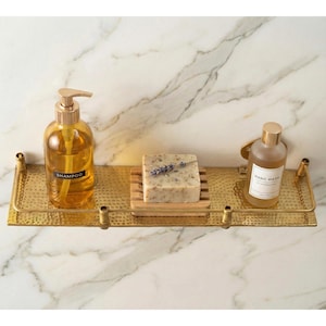 May include: A gold-colored metal bathroom shelf with a hammered texture. It holds a clear shampoo bottle, a wooden soap dish with a bar of soap, and a hand wash bottle. The shelf is mounted on a marble-patterned wall.