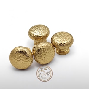 May include: Four gold cabinet knobs with a hammered texture. The knobs are round with a mushroom-shaped design and a small base. The image includes a logo with the text "PLUMBRA BRASS HARDWARE & FIXTURES".