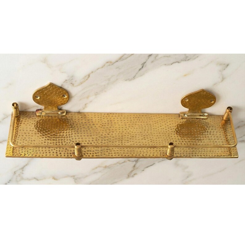 Hammered brass shelf installed in a bathroom, neatly organizing perfume bottles, a small plant, and rolled hand towels. The gold brass contrasts beautifully against the wall.