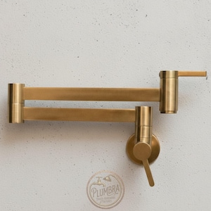 Puede incluir: Un grifo de llenado de ollas montado en la pared, de color latón. El grifo tiene un brazo de doble articulación que se extiende desde la pared. El cabezal y el mango del grifo son cilíndricos. El logotipo de la marca "Plumbra" es visible en la parte inferior.
