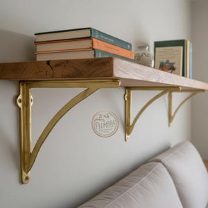 Puede incluir: Una estantería de madera con soportes de latón, montada en una pared blanca. La estantería contiene varios libros y un pequeño jarrón decorativo. Los soportes tienen un diseño curvo y están fijados a la pared con tornillos. La estantería es de madera clara.