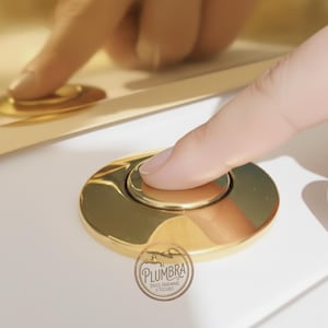 Op de afbeelding: Een close-up van een goudkleurige, cirkelvormige spoelknop die door een vinger wordt ingedrukt. De knop maakt deel uit van een sanitairarmatuur, met de merknaam "Plumbra" zichtbaar in een cirkelvormig logo.